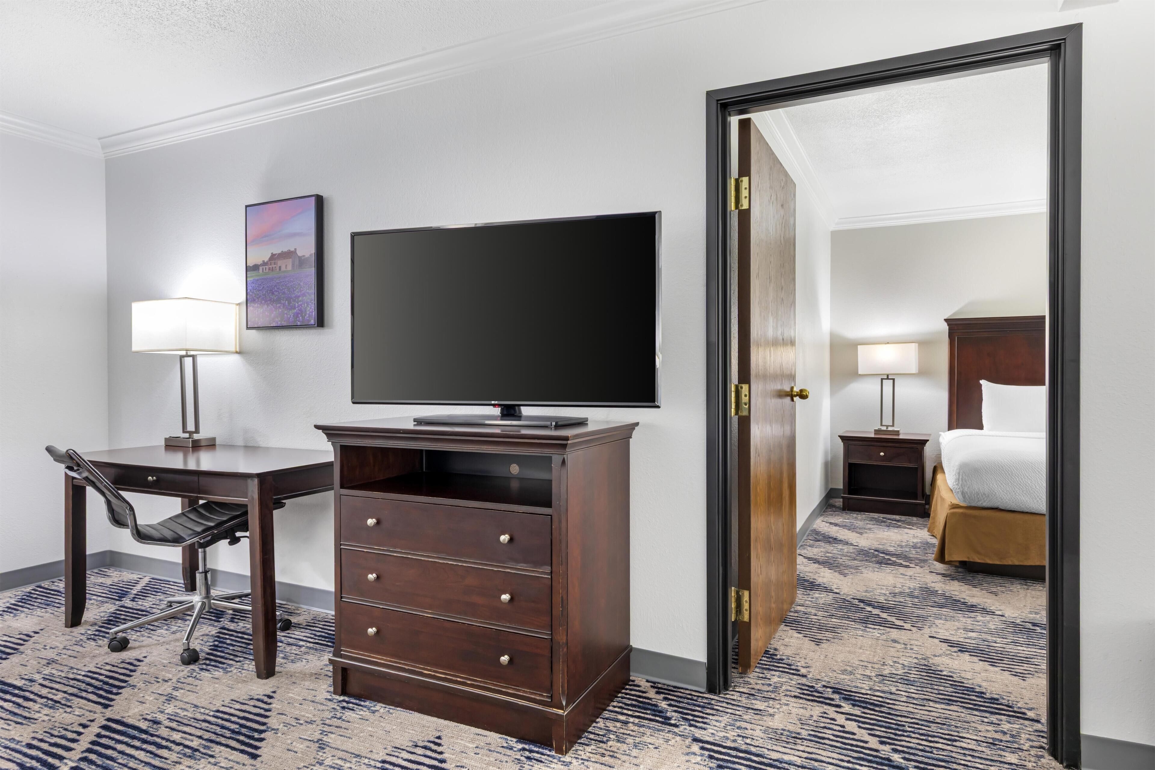 A hotel room with a flat-screen TV, a desk with a chair, a dresser, a bed, and a nightstand with a lamp.
