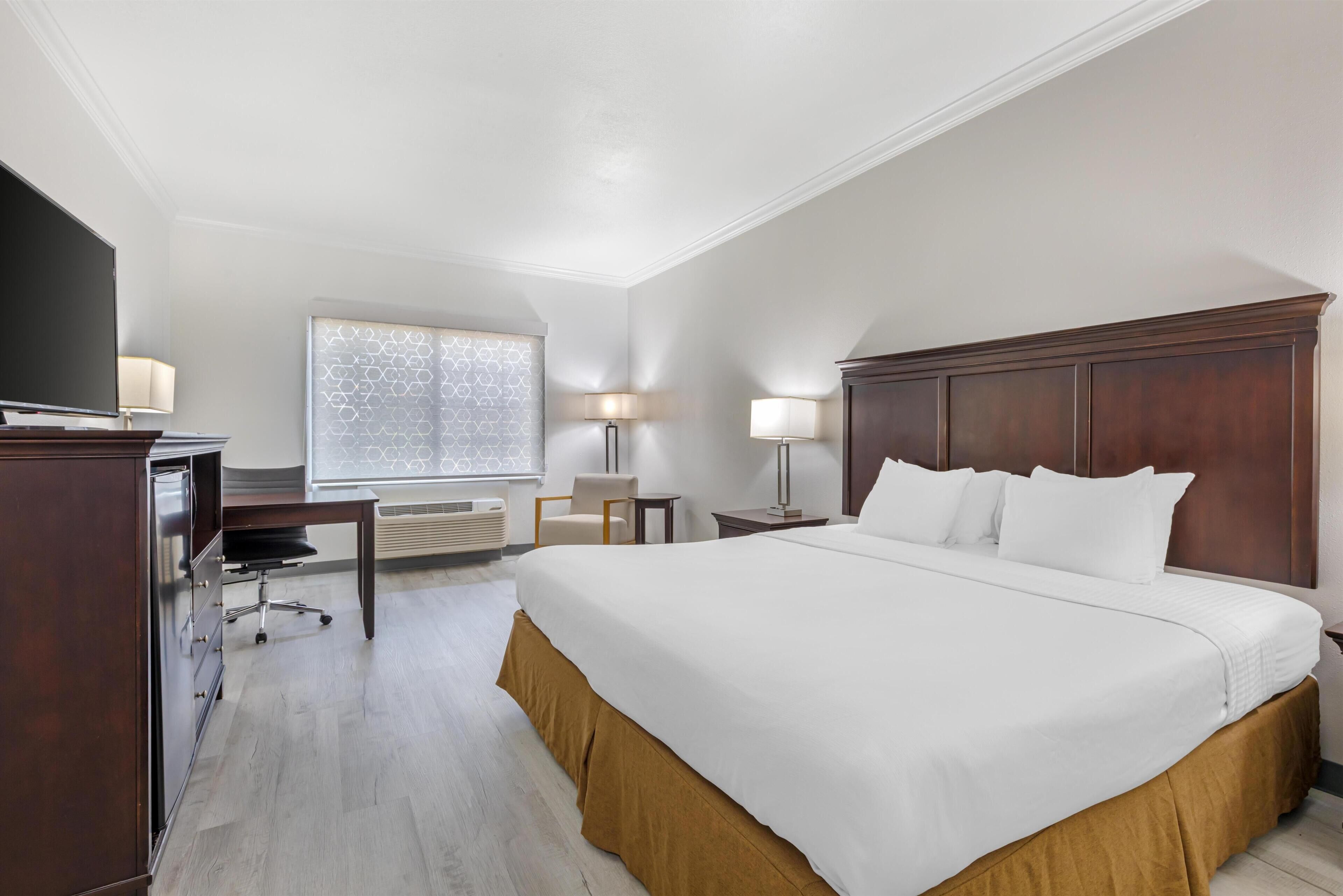 A hotel room with a large bed, a desk with a chair, a TV, a lamp, and a window with patterned curtains.