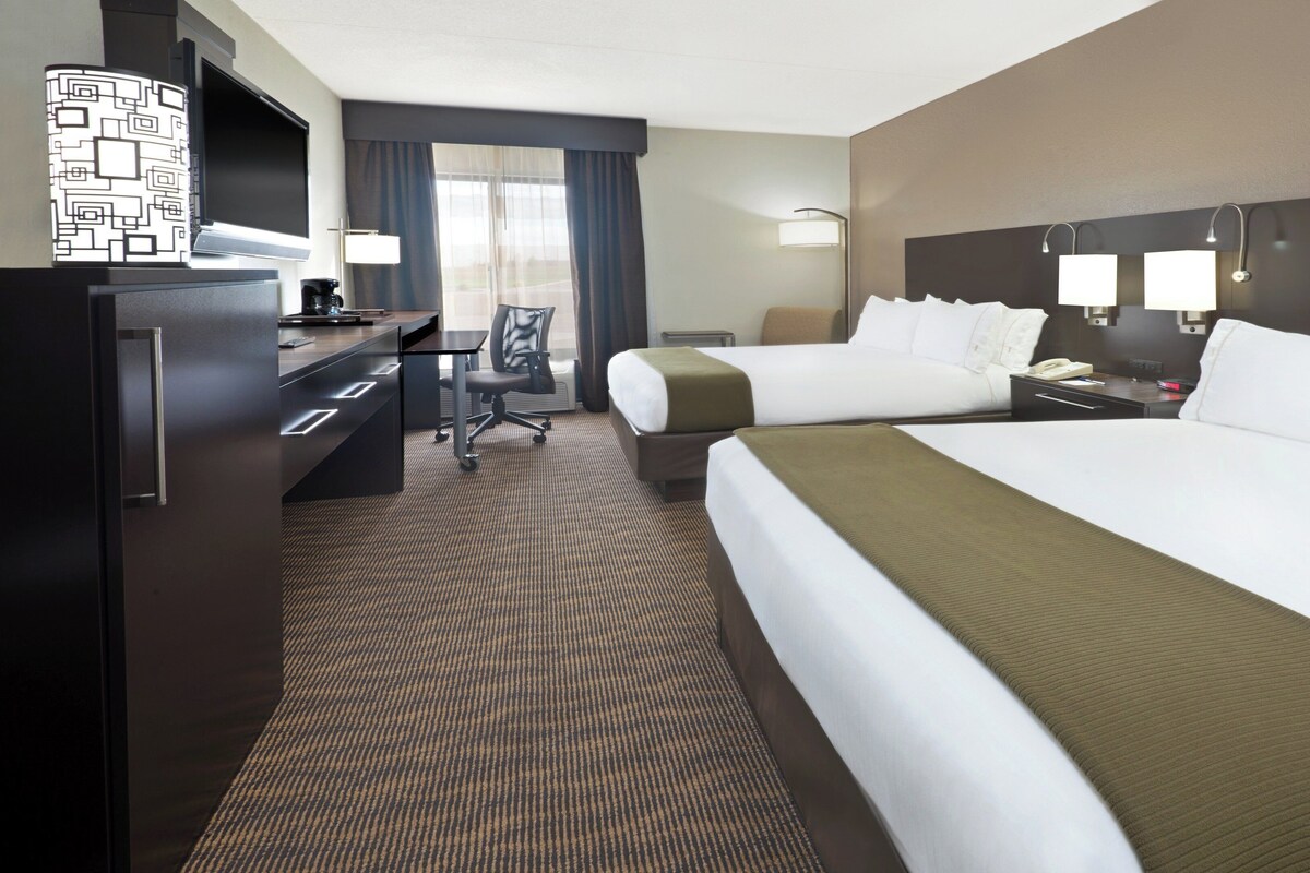 A hotel room with two beds, a desk, a chair, a minibar, and a large window with curtains.