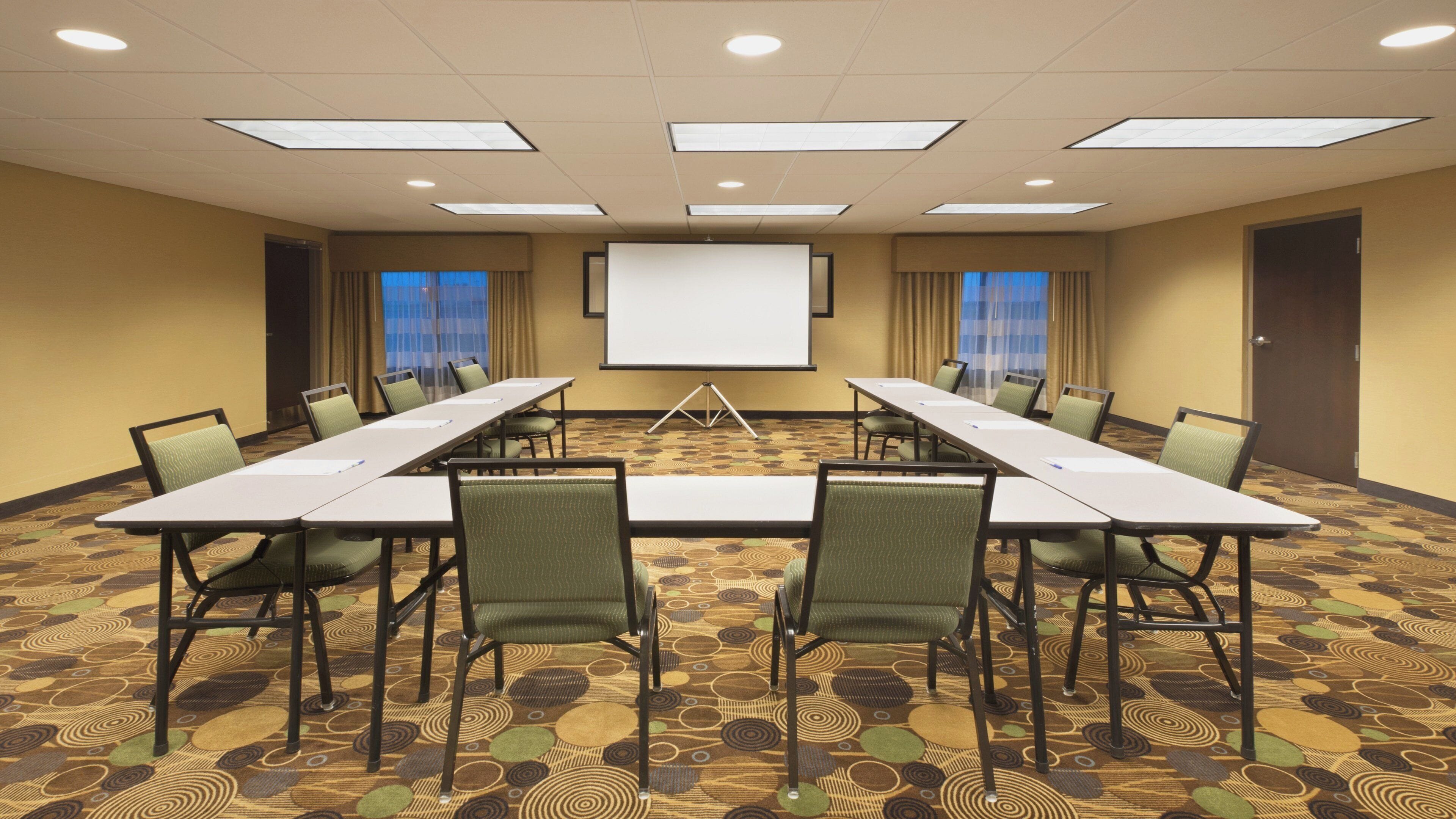 A conference room with a long table, chairs, a projector screen, and a carpeted floor.
