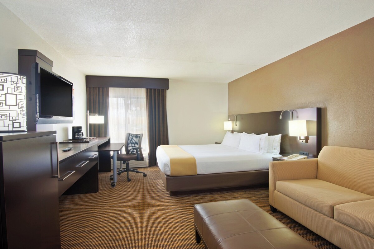 A hotel room with a bed, a desk with a TV, a chair, a sofa, and a carpeted floor.