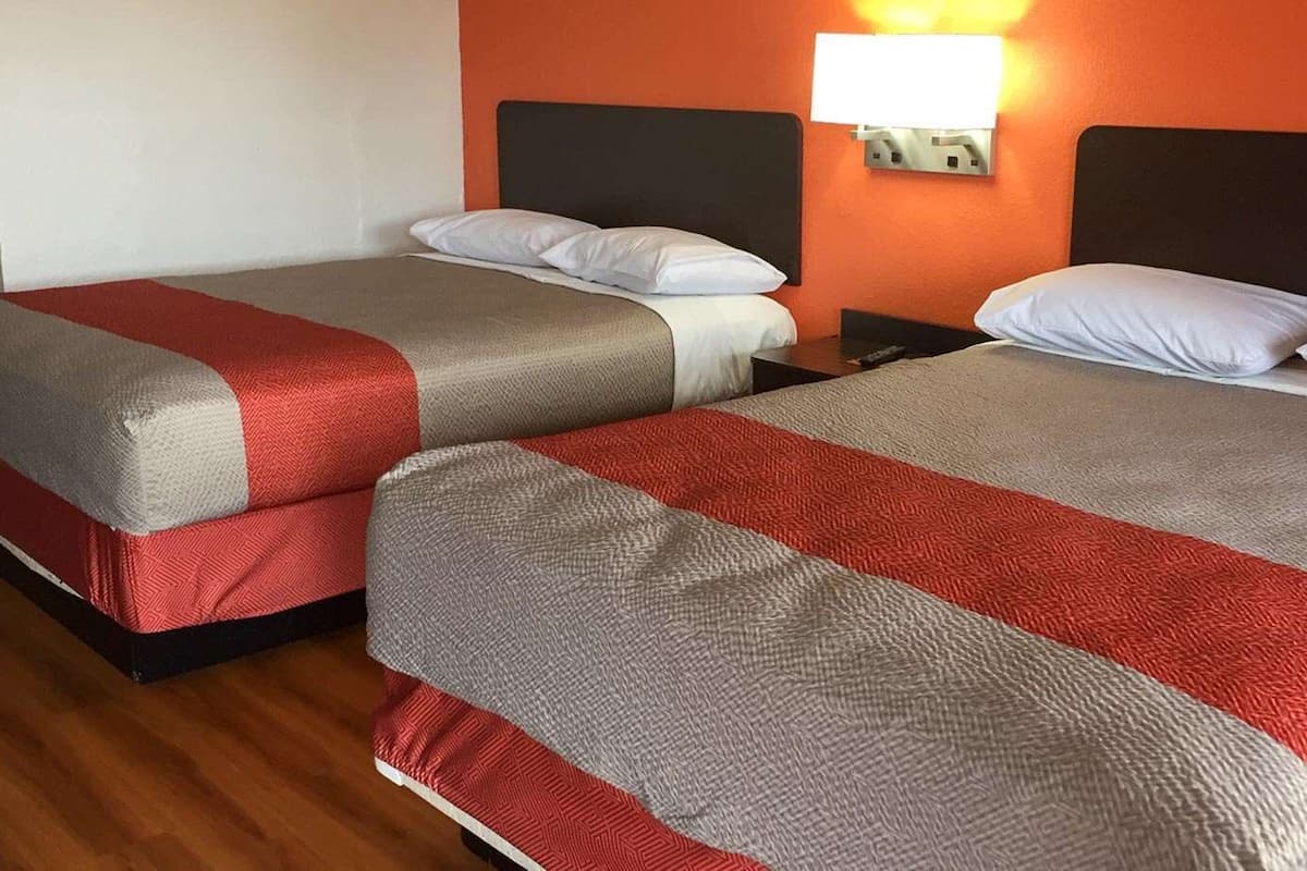 Quarto de hotel com duas camas, cada uma com colcha vermelha e cinza, e uma mesa de cabeceira com abajur entre elas.
