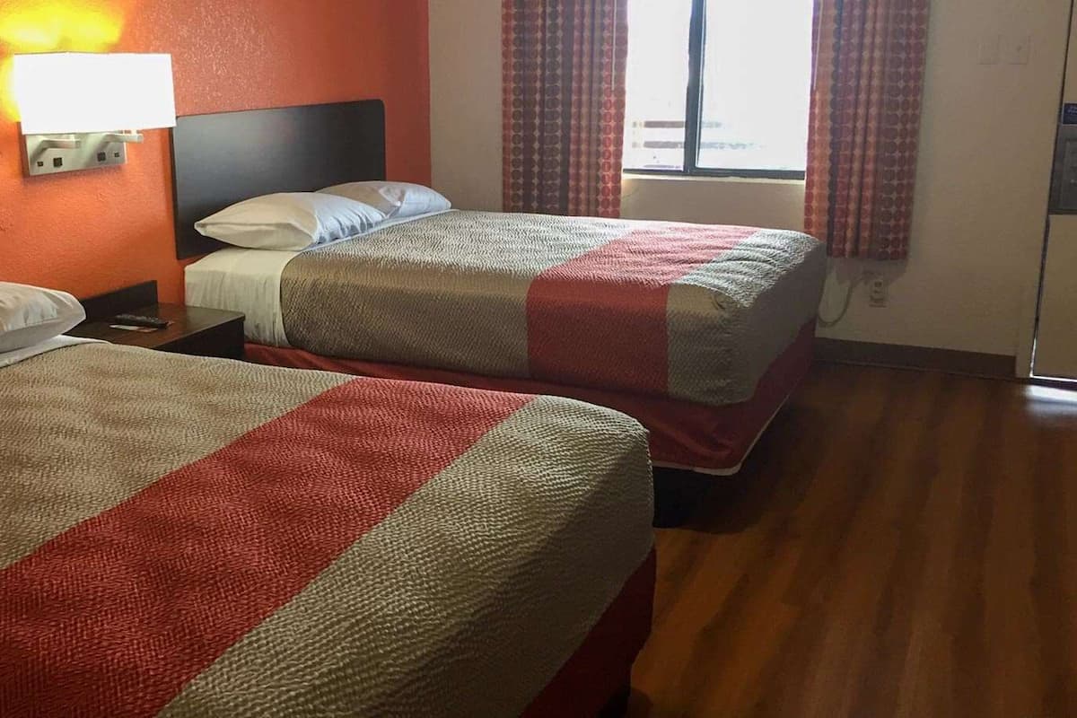 Duas camas de solteiro com roupa de cama vermelha e branca, uma mesa de cabeceira, uma luminária de parede e uma janela com cortinas.