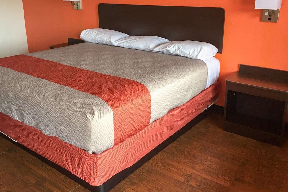 Uma cama com manta cinza e vermelha, travesseiro branco e mesa de cabeceira de madeira com abajur.