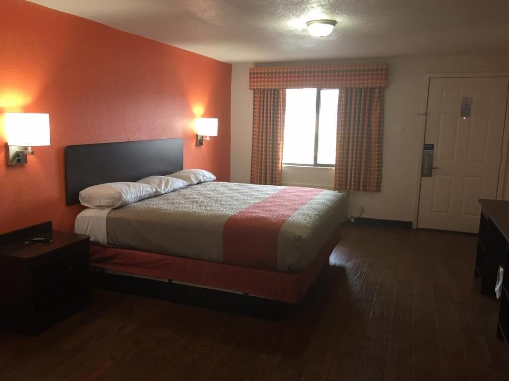 A hotel room with a bed, two bedside lamps, a window with curtains, and a door.