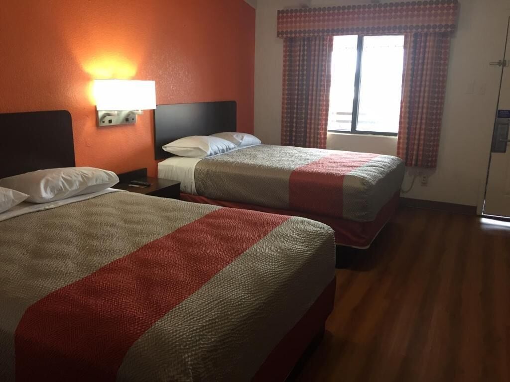 A hotel room with two beds, a nightstand, a lamp, and a window with curtains.