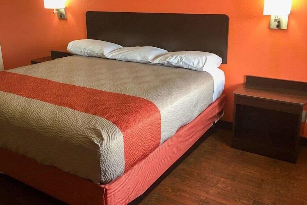Uma cama com manta vermelha e bege, duas almofadas e cabeceira de madeira. Há duas mesas de cabeceira com abajures, e as paredes são laranjas.