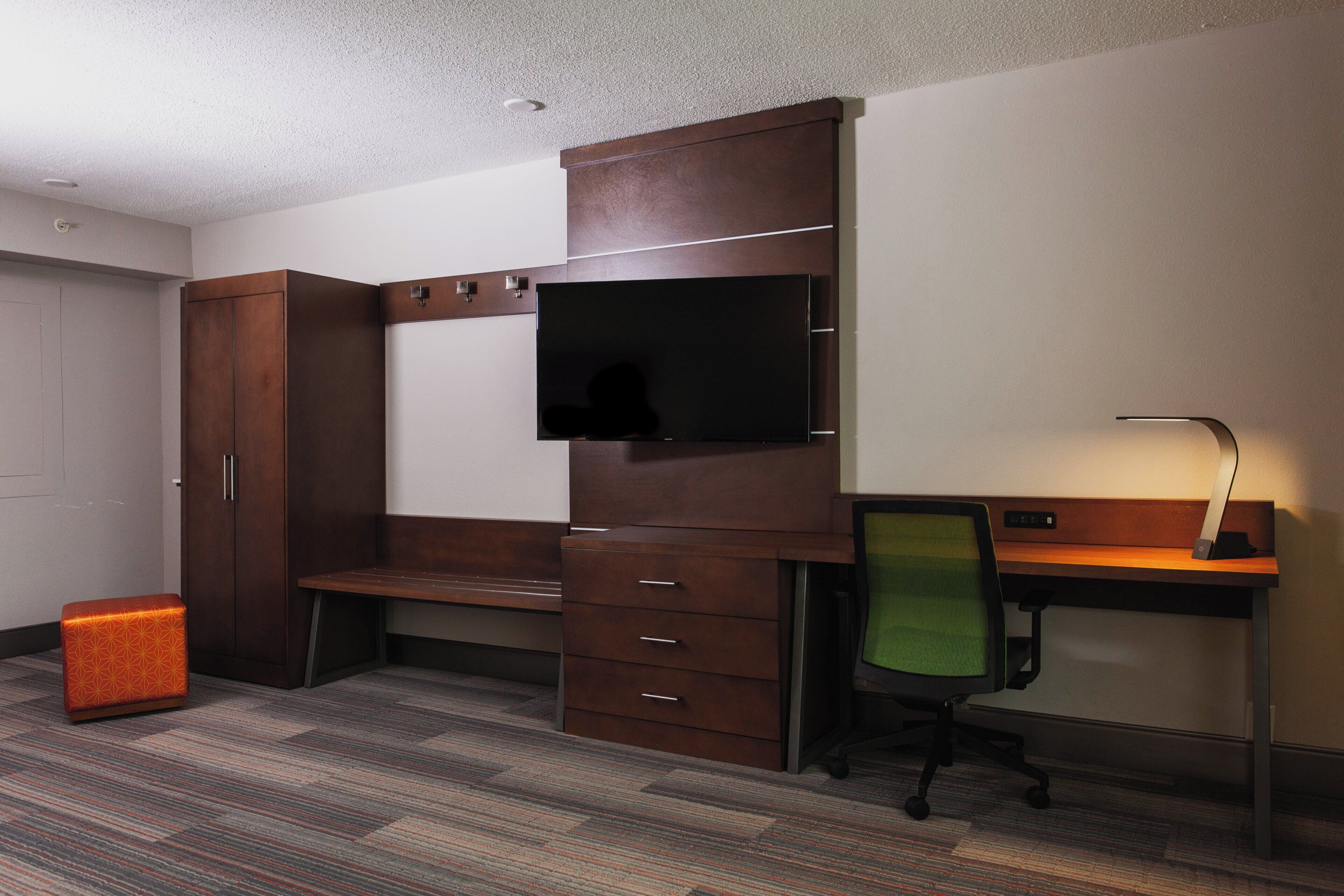 A hotel room with a desk, chair, television, wardrobe, and a trash bin.