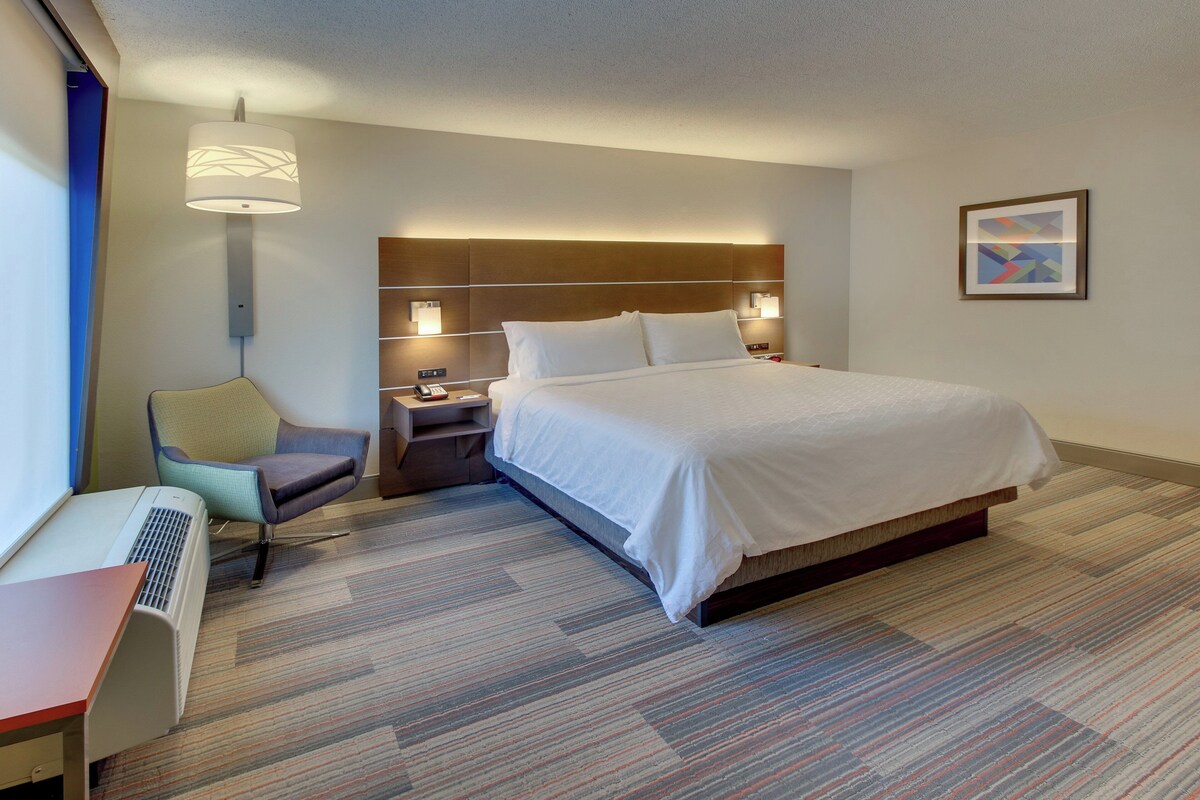 Ein Hotelzimmer mit Bett, Schreibtisch, Stuhl, Lampe und einem an der Wand hängenden Kunstwerk.