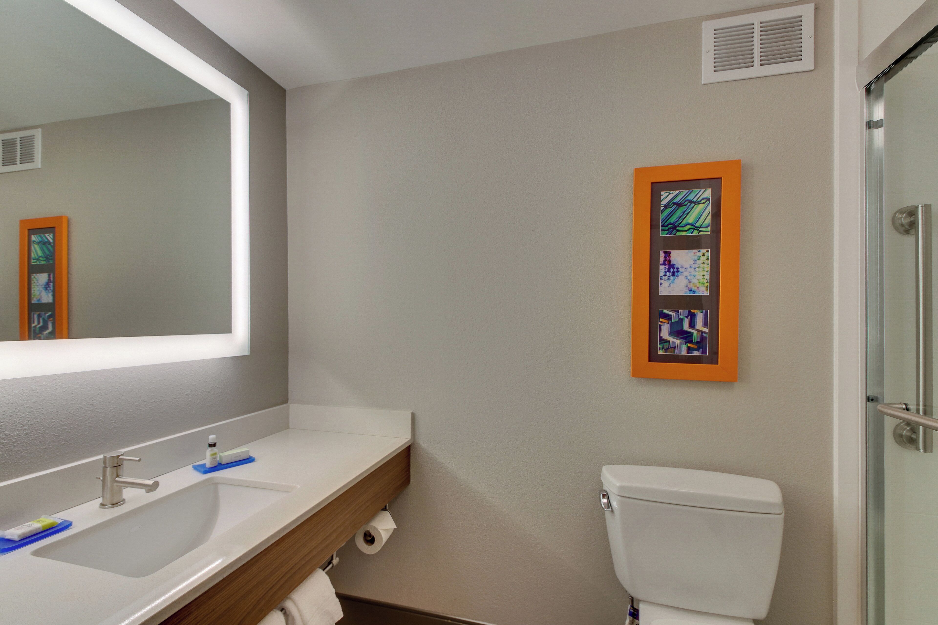 A bathroom with a sink, mirror, toilet, and a framed artwork on the wall.