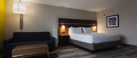 Quarto de hotel com cama, sofá, banqueta, mesa de cabeceira com abajur e quadro na parede.