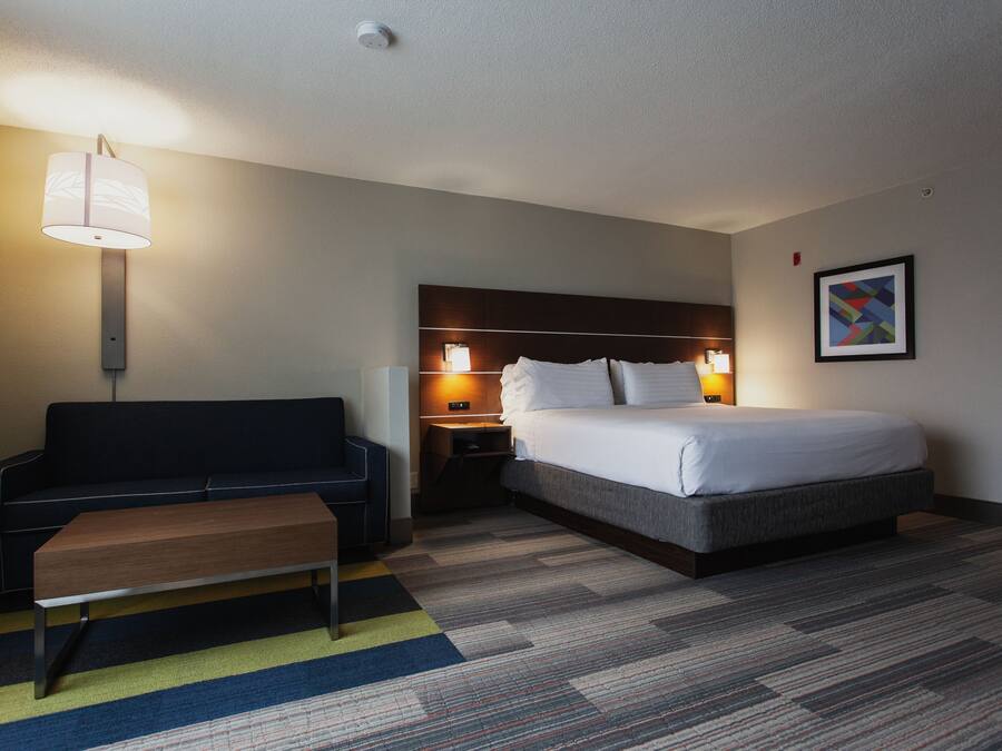 A hotel room with a bed, a sofa, a bench, a nightstand with a lamp, and a framed picture on the wall.