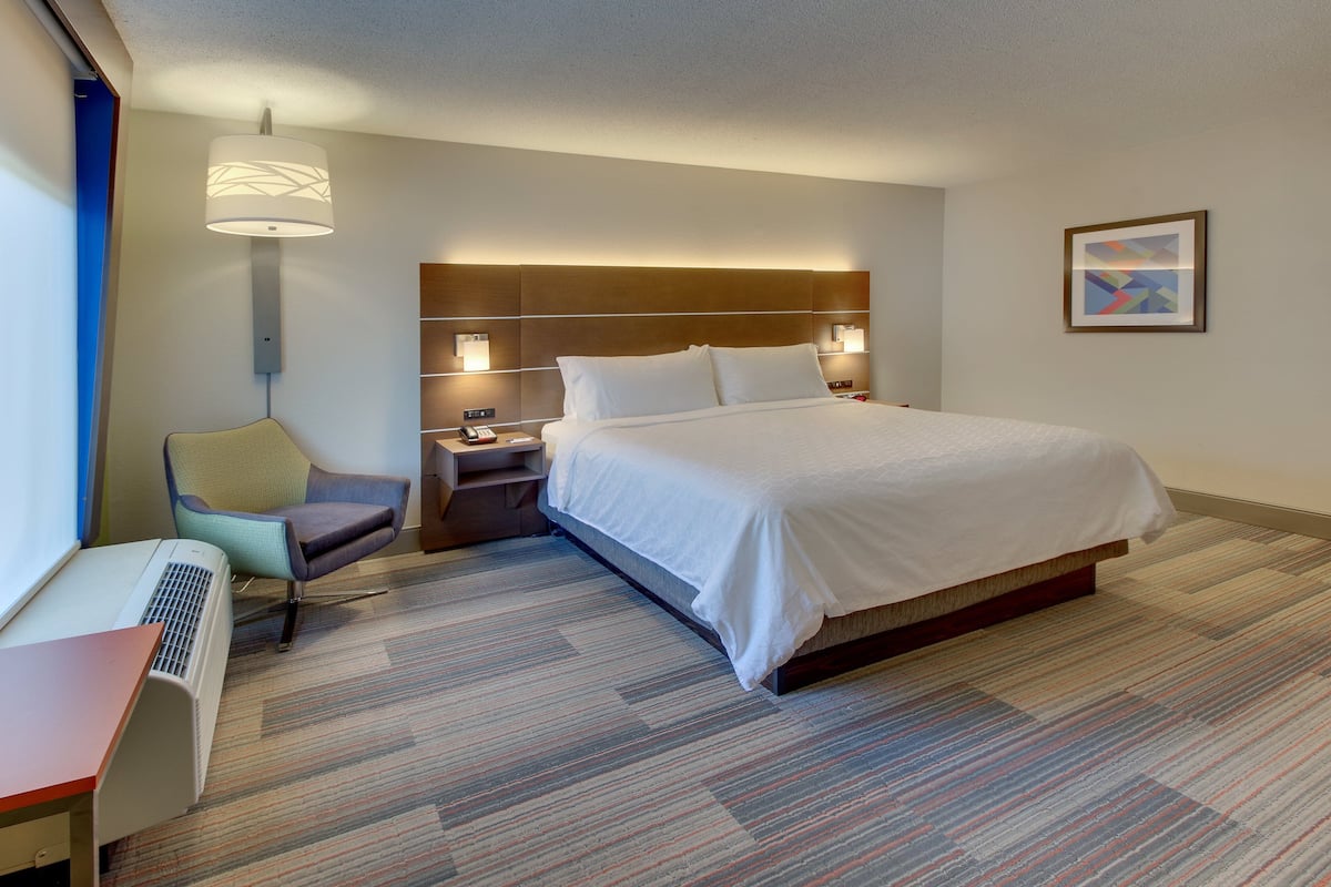 A hotel room with a large bed, a desk with a chair, a standing lamp, and a framed picture on the wall.