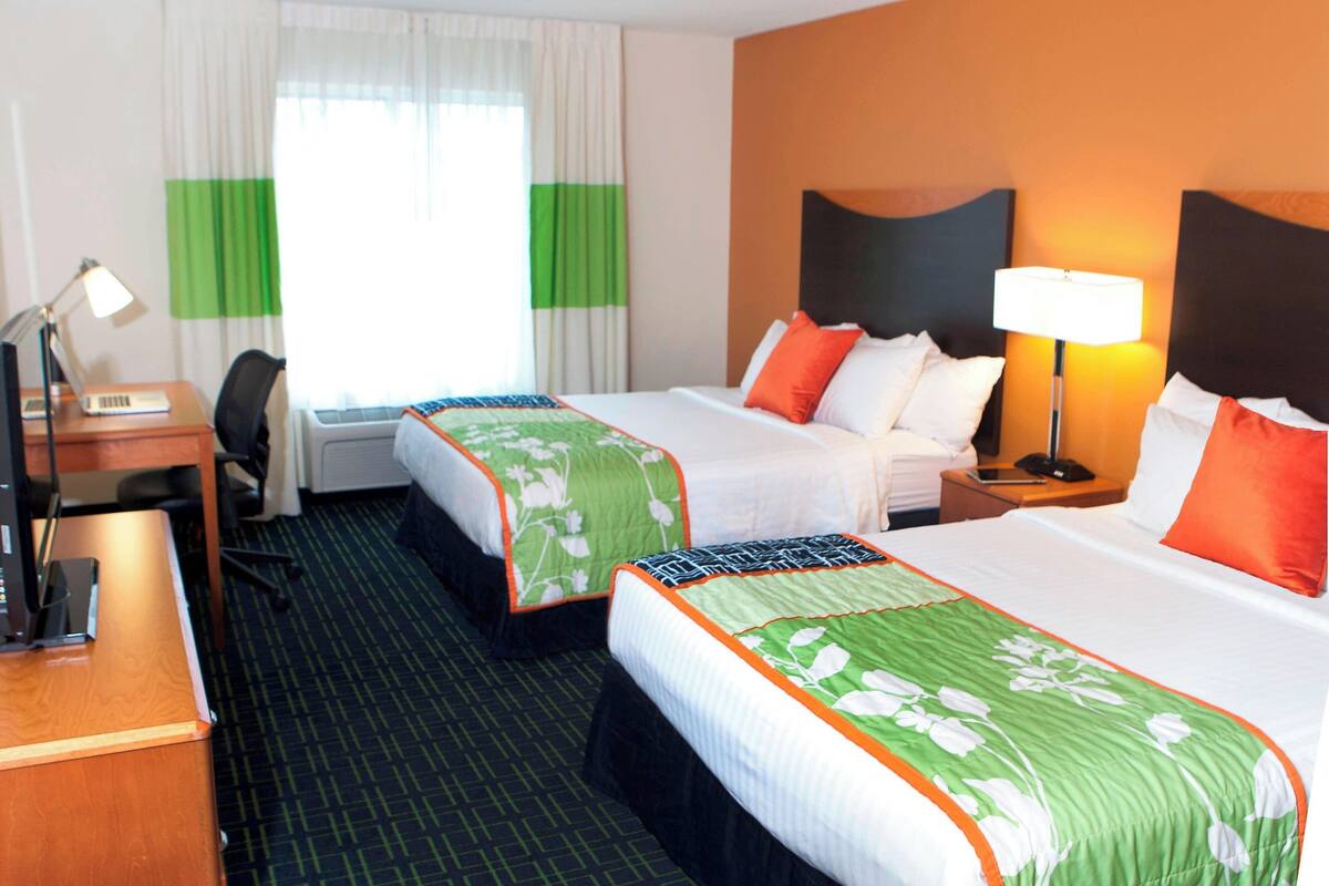 Une chambre d’hôtel avec deux lits, un bureau avec un ordinateur, une chaise, une lampe et une fen être avec des rideaux.
