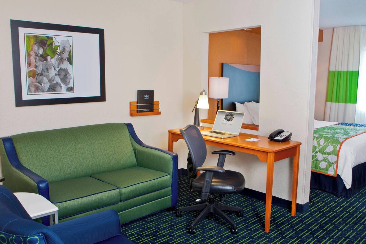 Une chambre d’hôtel avec un canapé vert, un bureau avec un ordinateur portable, une chaise, une table de chevet et un lit recouvert d’une couverture verte et blanche.