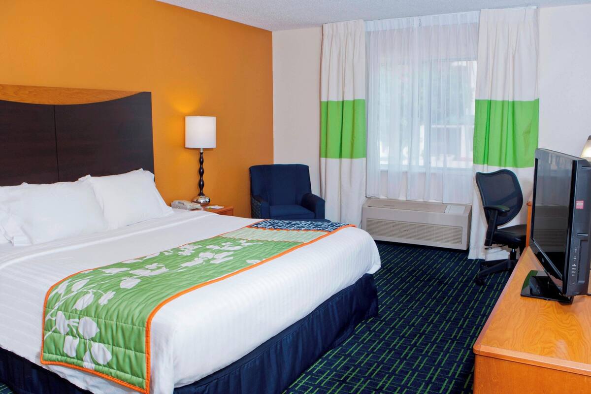 Une chambre d’hôtel avec un grand lit, un bureau avec un ordinateur, une chaise, une télévision et une fenêtre avec des rideaux.