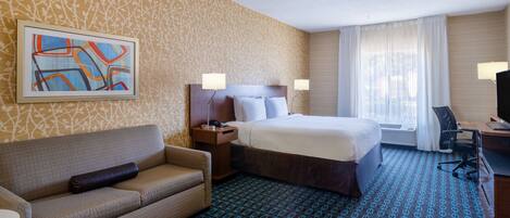 Una habitación de hotel con alfombra estampada, una cama con ropa blanca, un sofá, un escritorio con silla y un cuadro en la pared.