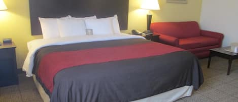 A hotel room with a large bed, a red armchair, a nightstand, and a lamp.