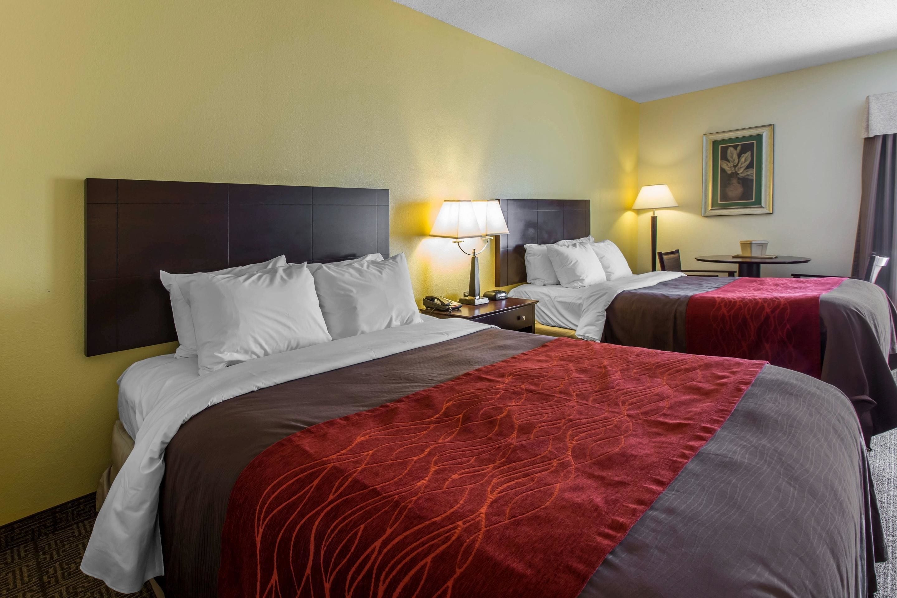 A hotel room with two beds, each with white bedding and a red accent. There are bedside tables with lamps, a small round table, and a framed picture on the wall.