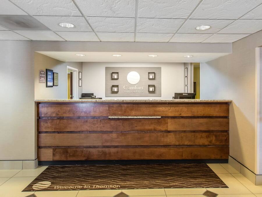 A hotel reception area with a wooden reception desk, a sign reading "Comfort Inns & Suites," and a welcome mat.
