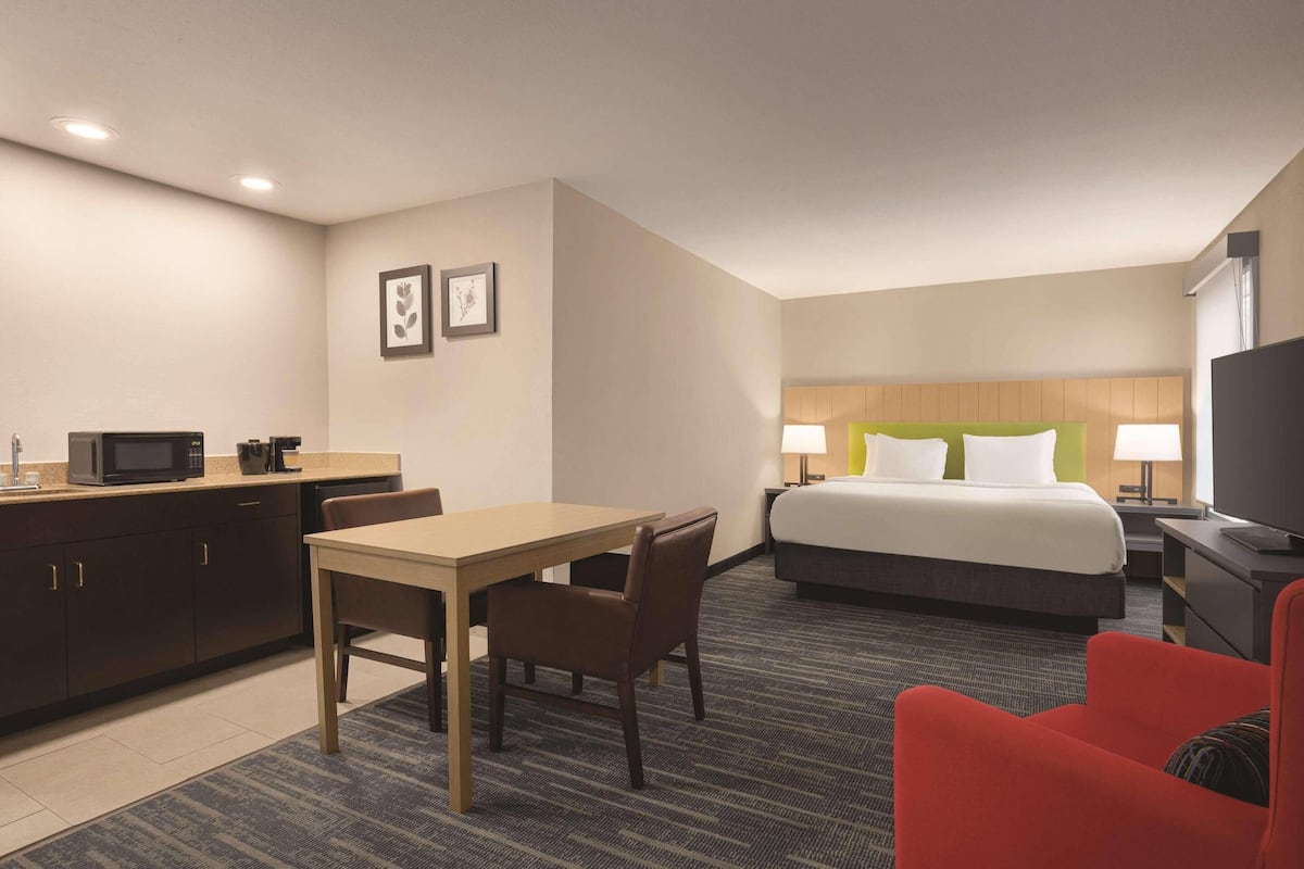 A hotel room with a bed, a desk with a microwave, a chair, a red armchair, and a TV.