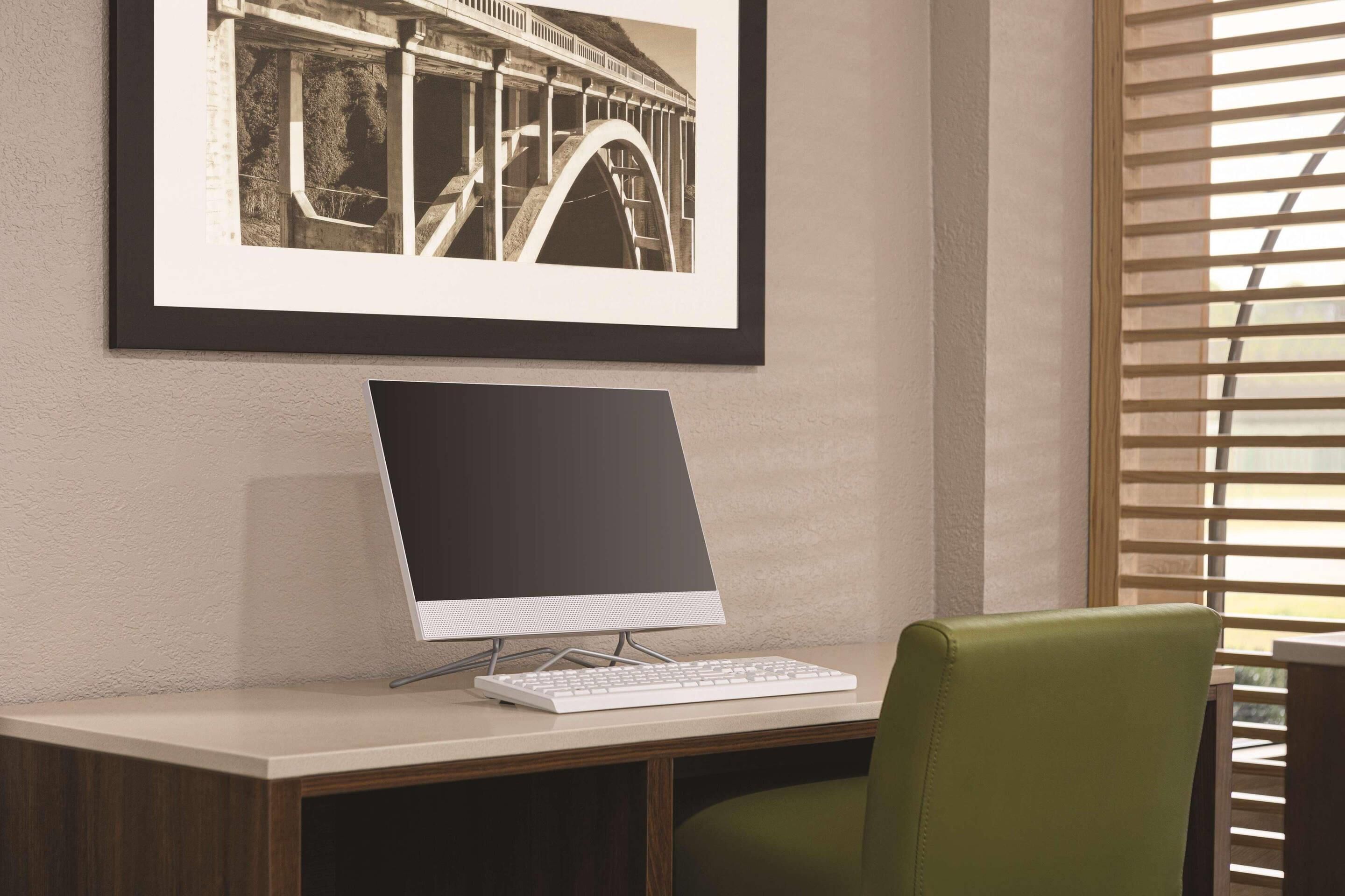 A desk with a computer monitor, keyboard, and a framed picture on the wall.