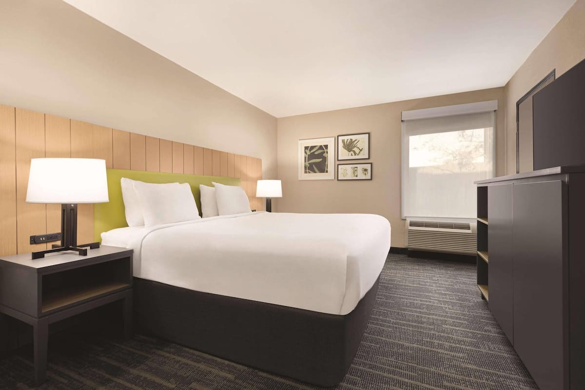 A hotel room with a bed, nightstands, a desk, a window with blinds, and wall art.