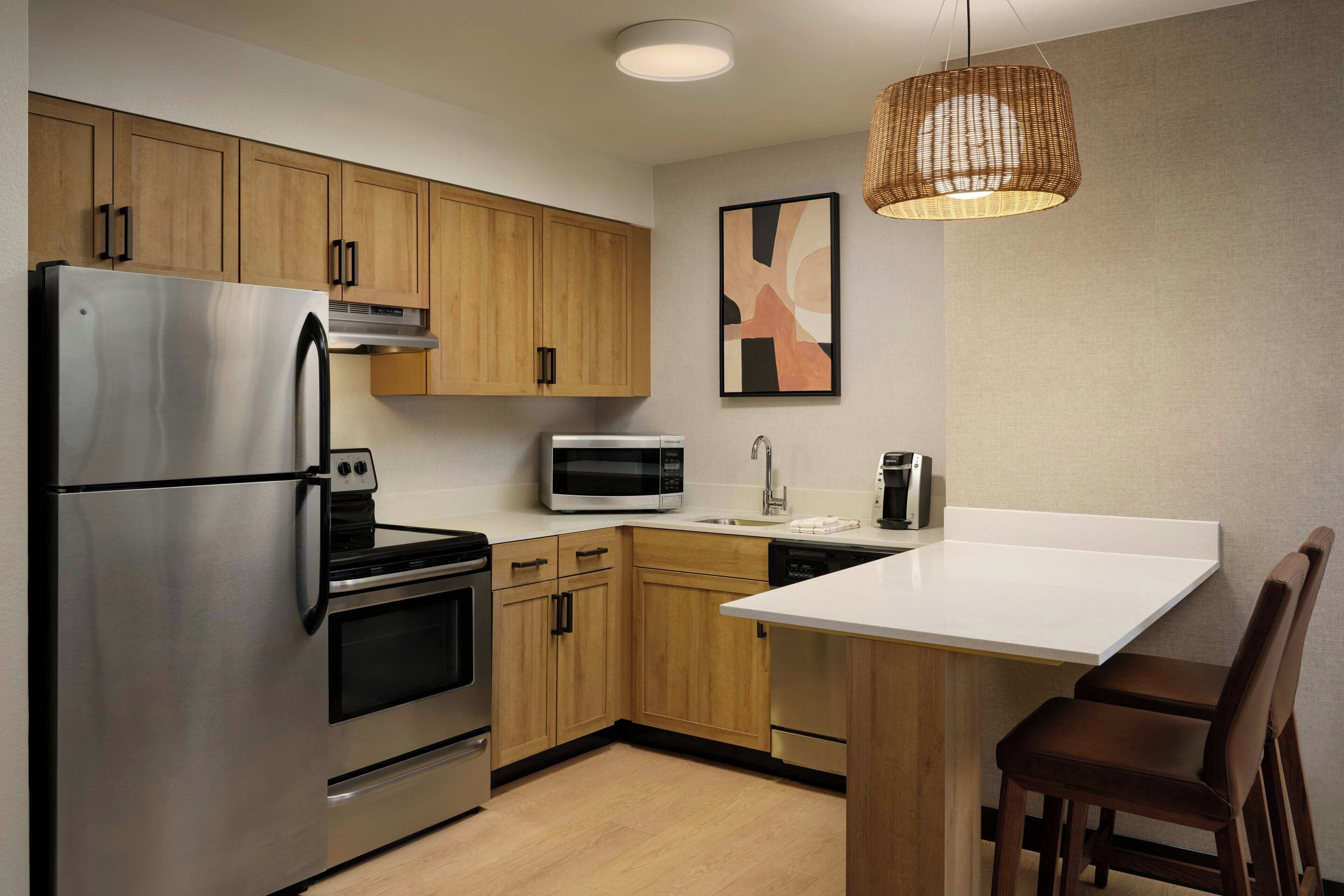 A modern kitchen with stainless steel appliances, wooden cabinets, a white countertop, and a hanging light fixture.