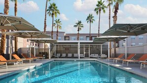 Una piscina con sillas de descanso y sombrillas, rodeada de palmeras y un edificio de diseño moderno.