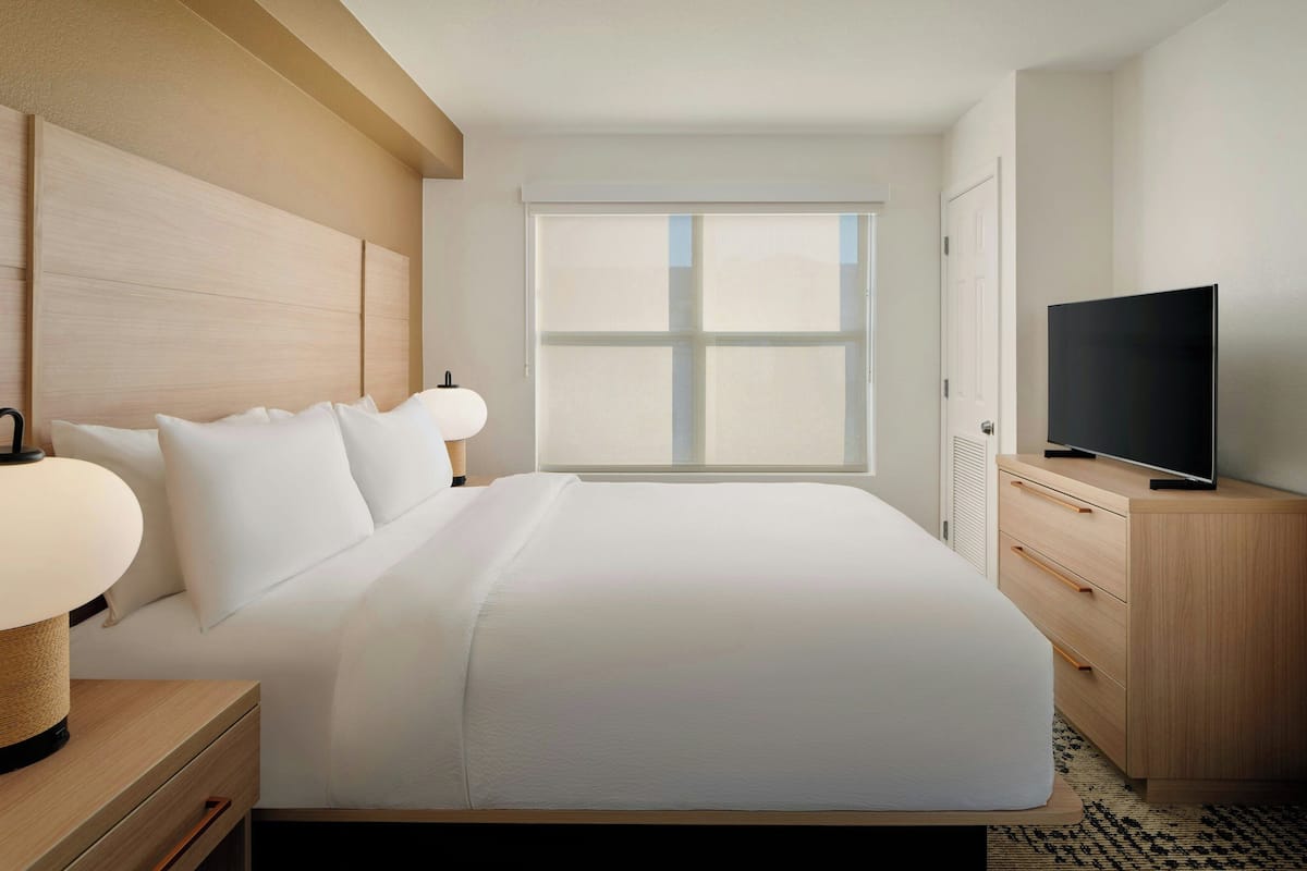 A hotel room with a large bed, a TV on a wooden cabinet, a window with blinds, and a bedside lamp.