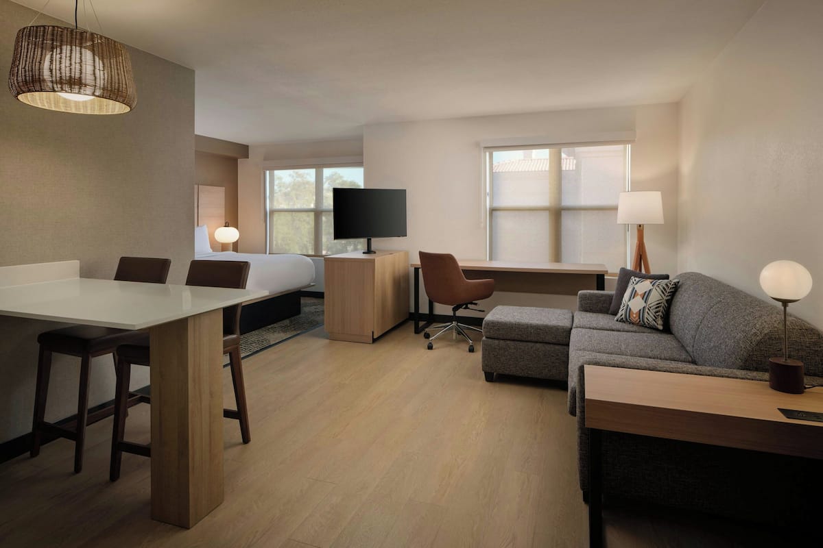 A modern hotel room with a bed, desk, office chair, sofa, and a dining table with chairs.