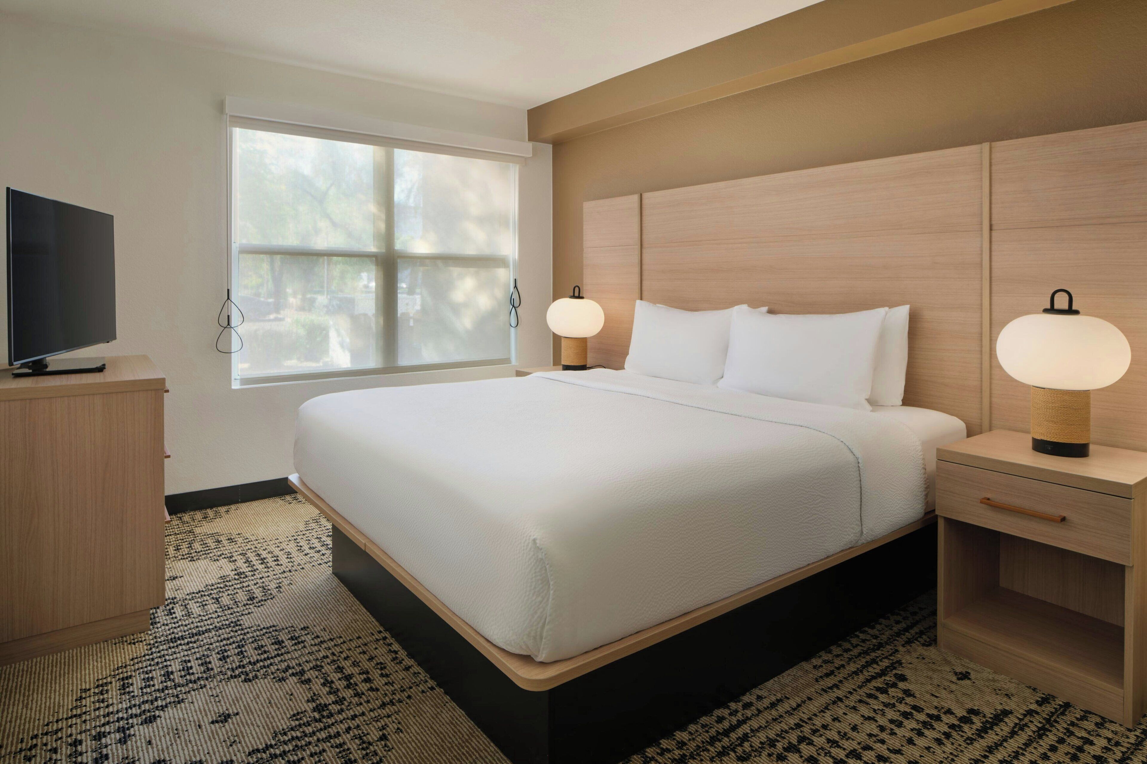 Une chambre d’hôtel bien aménagée, avec un grand lit, des lampes de chevet, une télévision et une fenêtre équipée de stores.