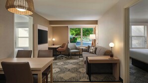 Habitación de hotel moderna con zona de comedor, sofá, televisor y un amplio ventanal con vistas a zonas verdes.