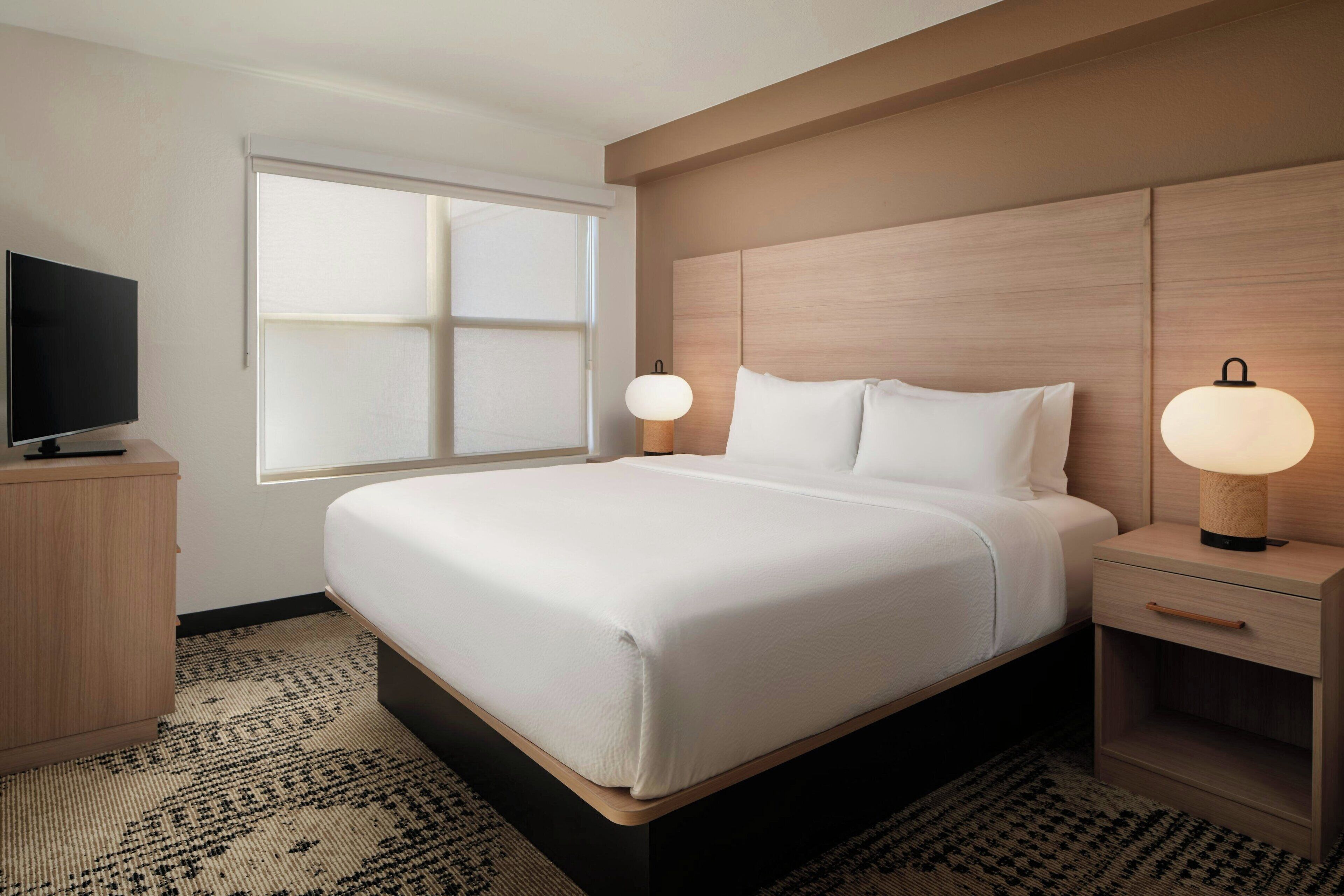 Een moderne hotelkamer met een groot bed, bedlampen, een flatscreen televisie en een raam met zonwering.