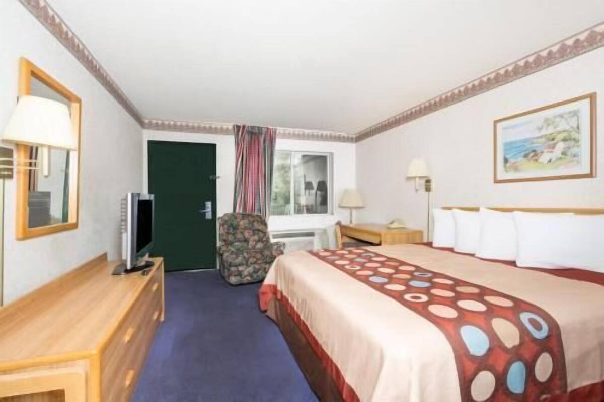A hotel room with a bed, a desk, a chair, a TV, a window with curtains, and a painting on the wall.