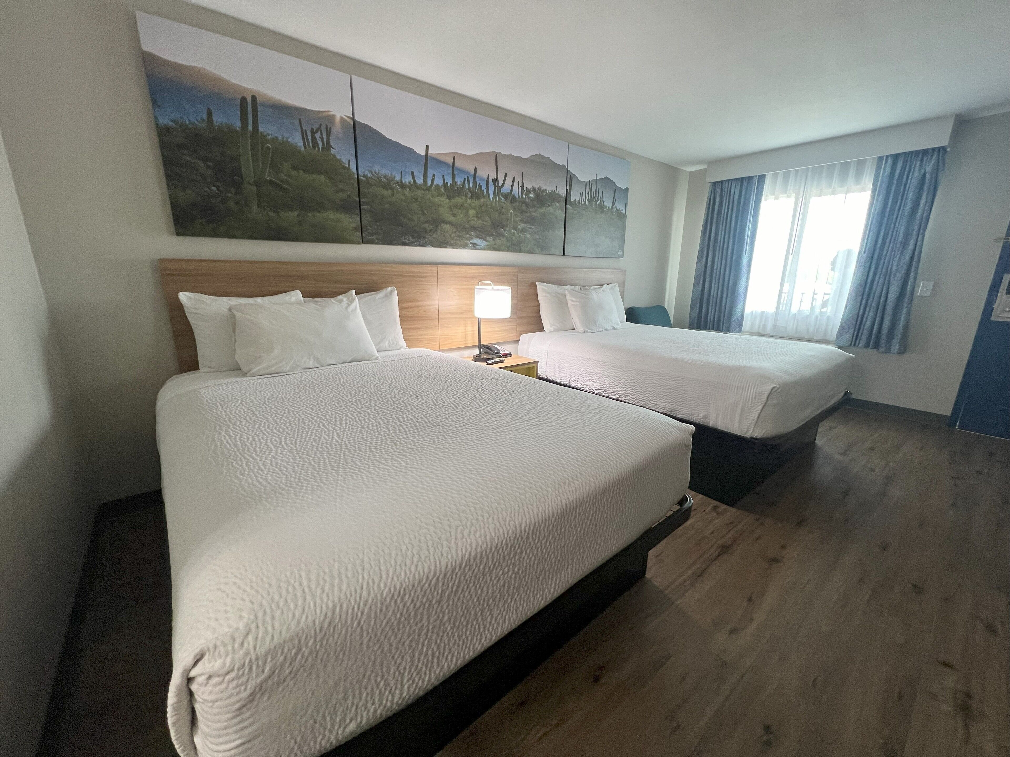 A hotel room with two beds, a wooden headboard, a nightstand with a lamp, and a large landscape painting on the wall.