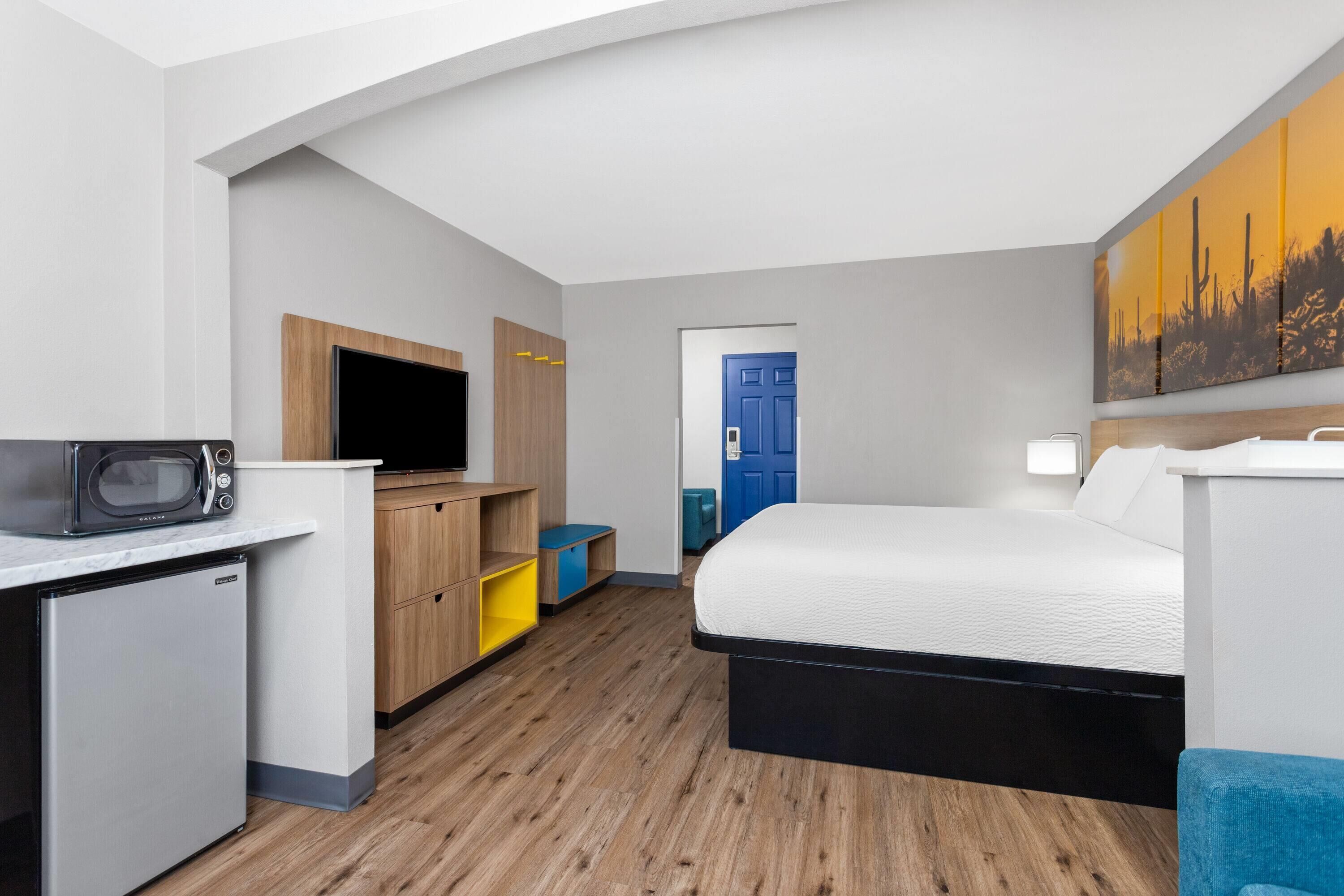 A compact hotel room with a kitchenette, a bed, a TV, and a painting on the wall.