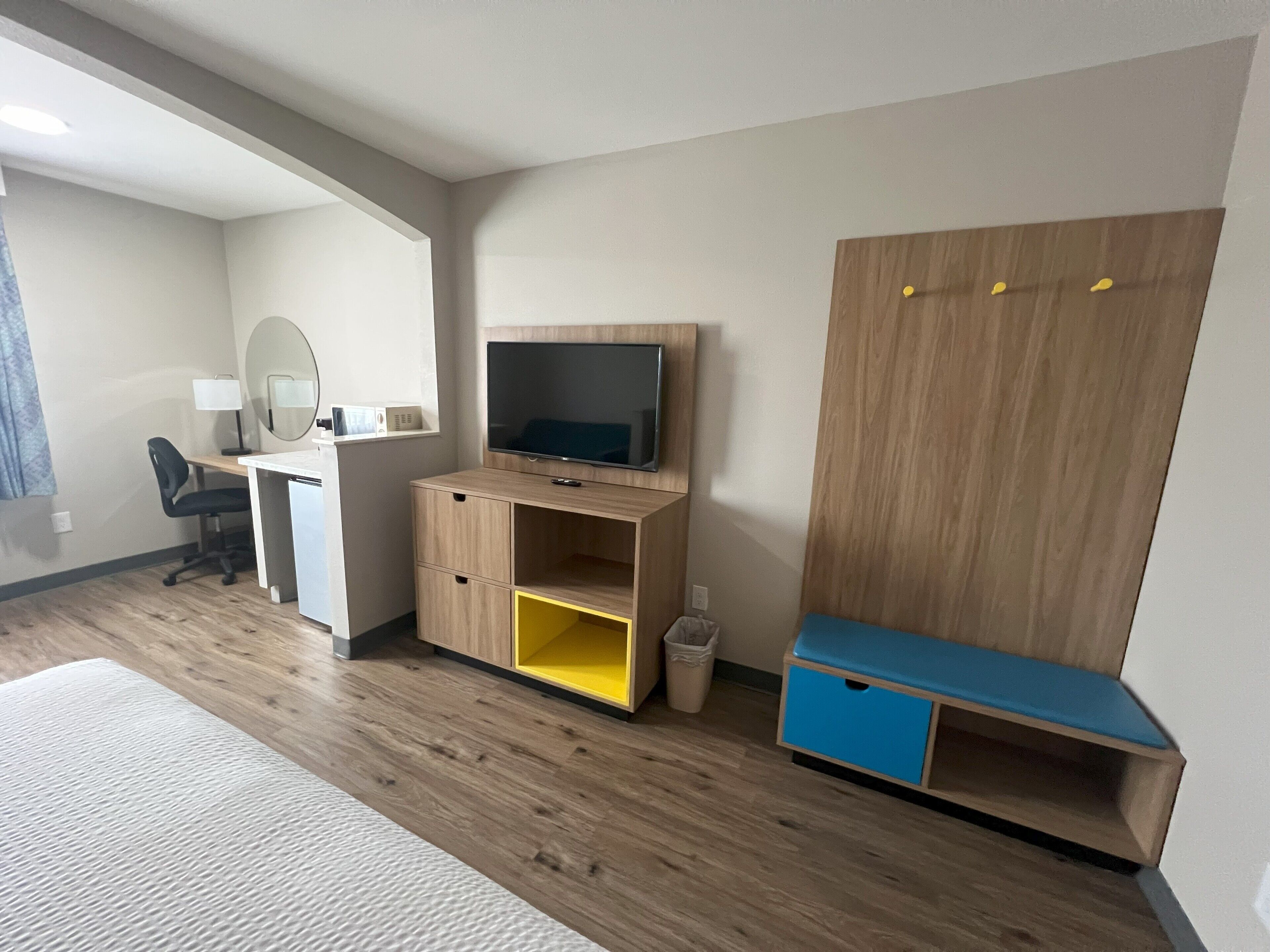 A modern hotel room with a wooden desk, a television, a blue bench, and a large wardrobe.