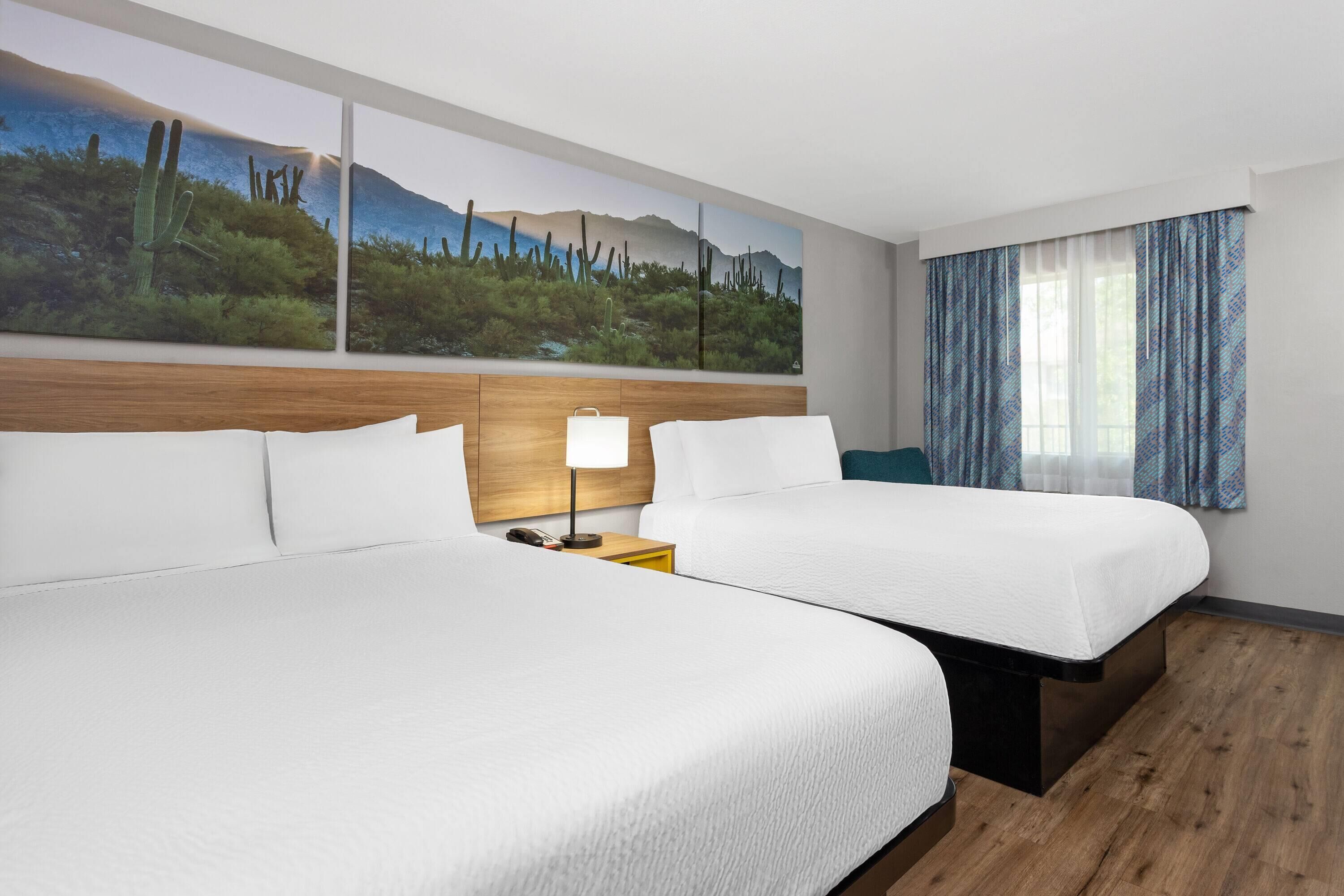 Habitación de hotel con dos camas, una mesita de noche con lámpara, un ventanal con cortinas y un cuadro de un paisaje desértico en la pared.