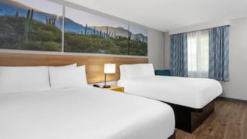 Habitación de hotel con dos camas, una mesita de noche con lámpara, un ventanal con cortinas y un cuadro de un paisaje desértico en la pared.