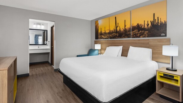 A hotel room with a large bed, a wooden headboard, a nightstand with a lamp, a painting of a desert landscape, and a bathroom visible through an open door.