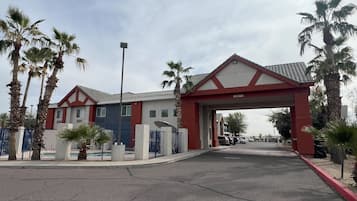 A gated entrance with a red archway leading to a building with a tiled roof and a blue door. Palm trees line the driveway.