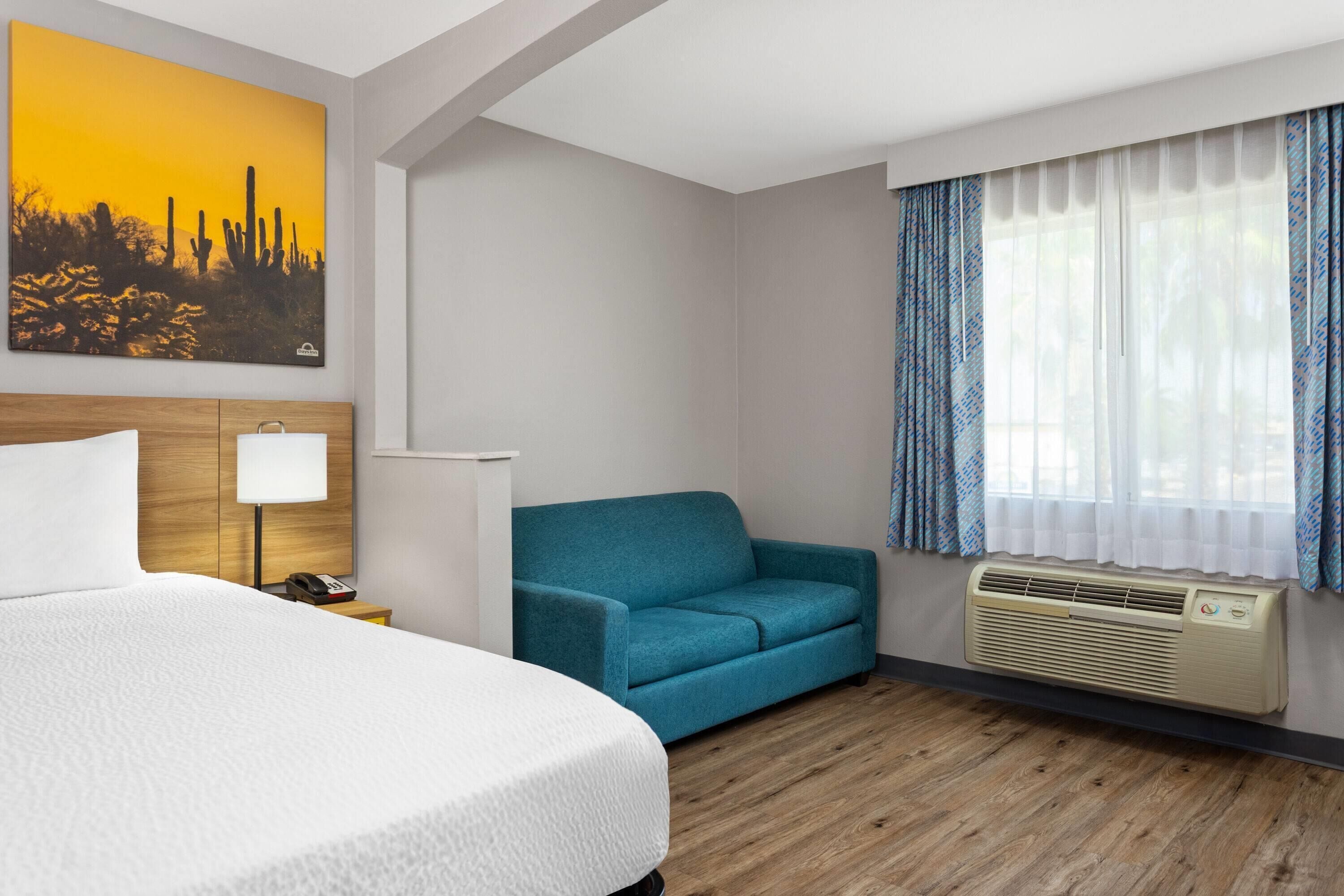 A hotel room with a bed, a blue sofa, a wall-mounted air conditioner, and a painting of a desert landscape.