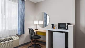 Een compacte hotelkamer met een bureau, stoel, magnetron, minibar en een groot raam met dunne gordijnen.