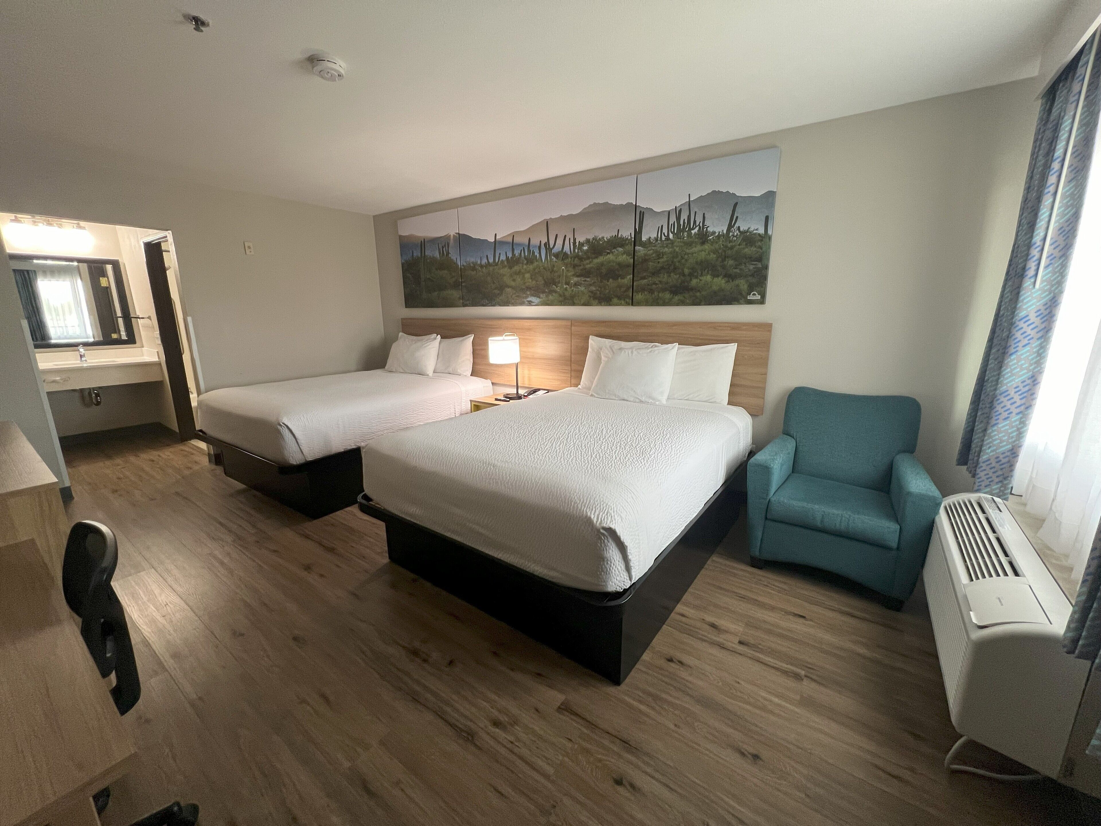 Een hotelkamer met twee bedden, een bureau, een stoel en een schilderij van een woestijnlandschap.