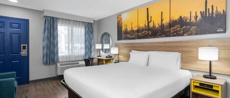 A hotel room with a large bed, a desk, a chair, a blue door, and a painting of cacti.