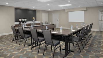 Uma sala de conferência com uma mesa comprida, cadeiras, tela de projeção e um espaço para café.