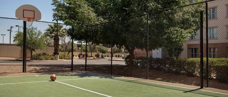 Una cancha de baloncesto al aire libre con aro, rodeada por una cerca de malla y árboles.