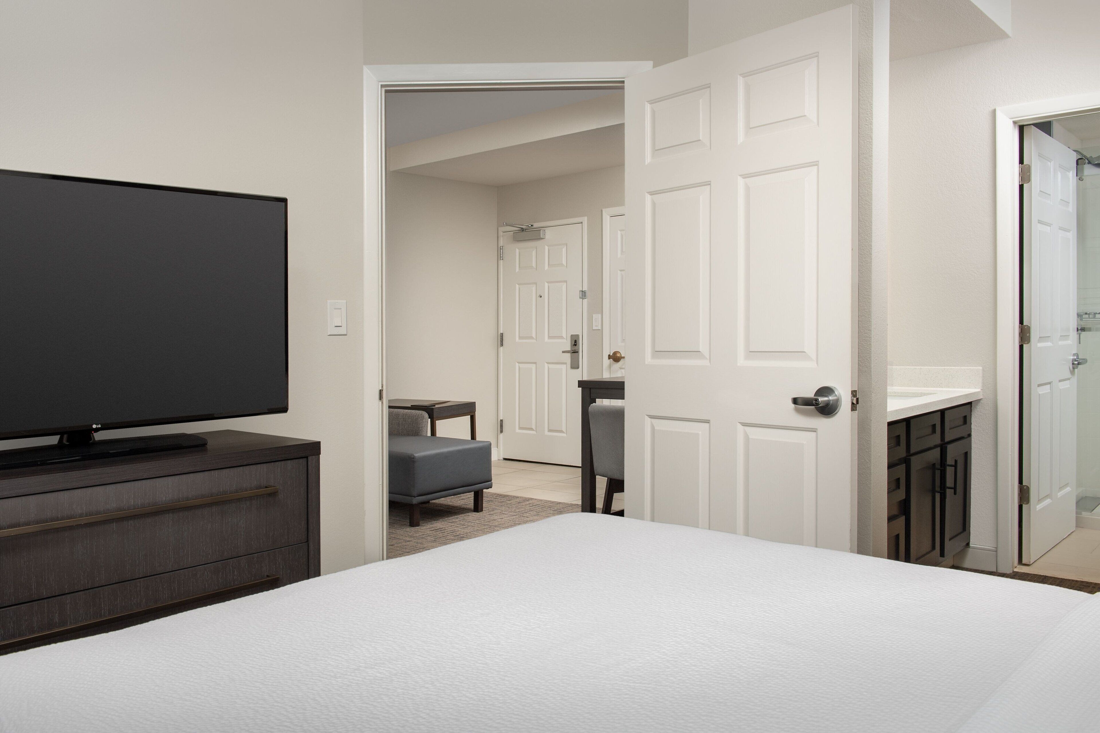 Quarto com cama, televisão em uma cômoda, cadeira e porta que dá para um banheiro.