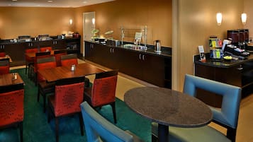 A hotel dining area with multiple tables and chairs, a buffet counter with various food items, and a coffee station.